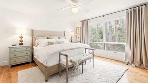 Un dormitorio con cama, mesitas de noche, un banco, ventilador de techo y una ventana grande con cortinas.