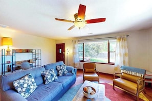 Ein Wohnzimmer mit einem blauen Sofa, einem braunen Ledersessel, einem Deckenventilator und einem Fenster mit Vorhängen.