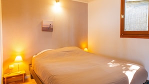 A single bed with a wooden frame, two bedside lamps, a small bedside table, a wall-mounted picture, and a window with a view of a wooden fence.