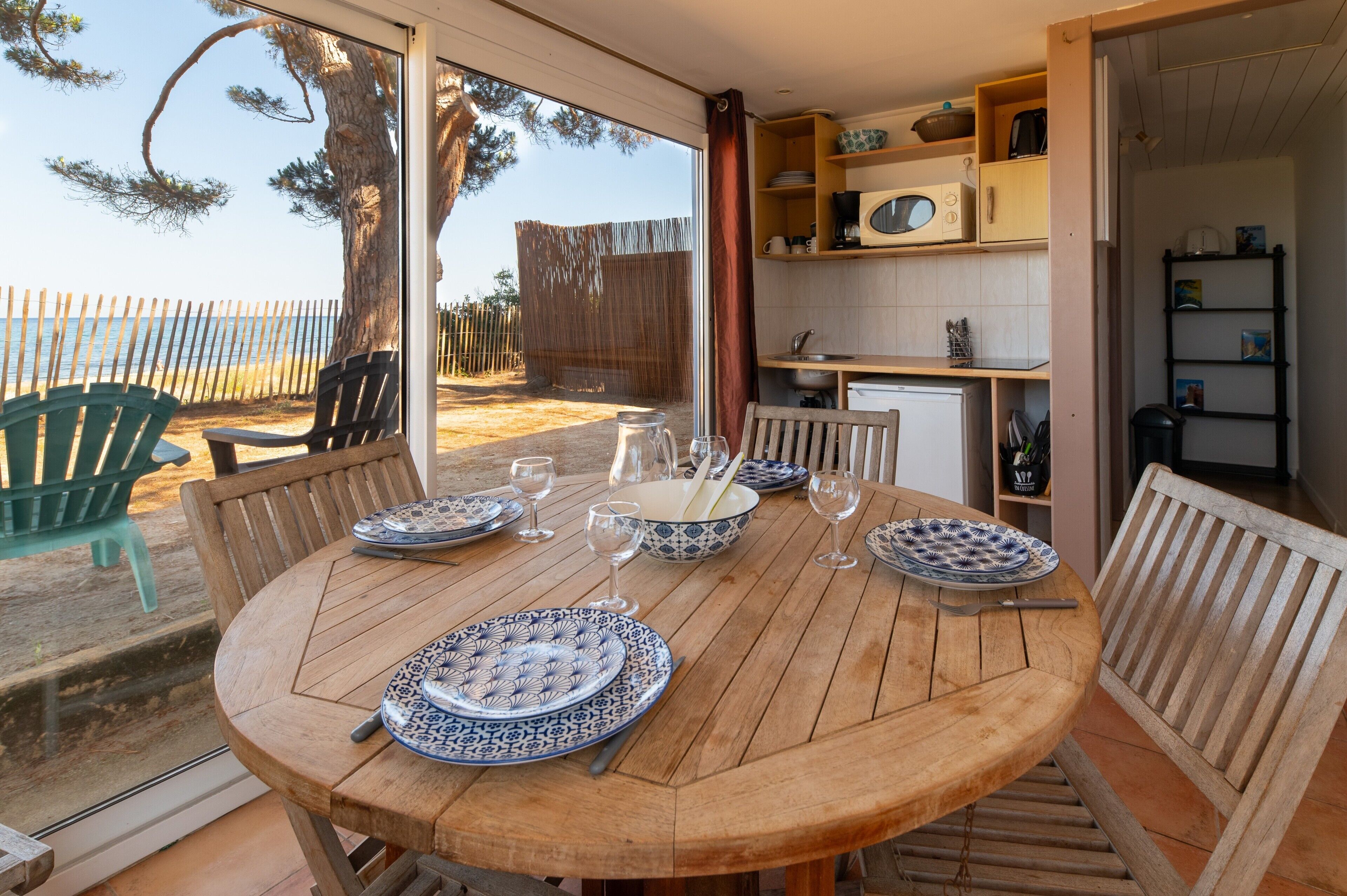Une table de salle à manger en bois, mise avec des assiettes, des verres et des couverts, avec vue sur la plage et un coin cuisine comprenant des placards et un four à micro-ondes.