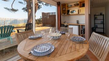 A wooden dining table set with plates, glasses, and cutlery, with a view of the beach and a kitchen area with cabinets and a microwave.