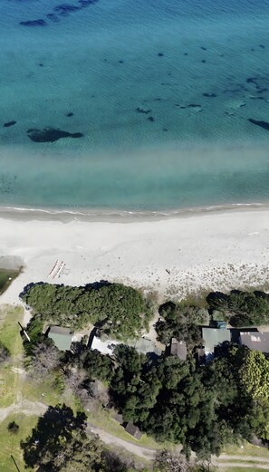 An aerial view of a beach with clear water, a sandy shore, and a cluster of buildings surrounded by dense greenery.