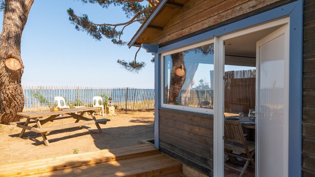 A wooden deck with a picnic table and chairs, a covered seating area with a table and chairs, and a view of the beach through a fence.