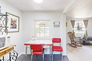 Un coin repas avec une table blanche, des chaises rouges, un fauteuil à bascule en bois, un four à micro-ondes et un tableau encadré au mur.