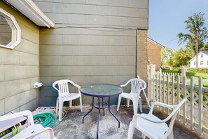 Un espace patio avec une table ronde et quatre chaises en plastique blanches, un tuyau d’arrosage et une clôture blanche.
