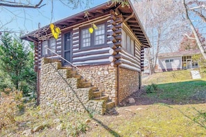 Une cabane en rondins avec un escalier en pierre menant à l’entrée, entourée d’arbres et avec un petit abri de jardin en arrière-plan.