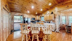 A wooden cabin interior with a kitchen, bar seating, and a view through the door.