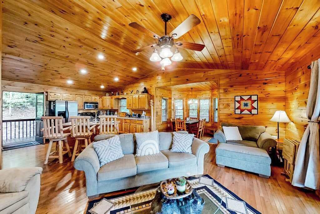 L’intérieur d’une cabane est doté d’un plafond en bois, d’un coin repas, d’un salon avec un canapé et un pouf, et d’une cuisine avec des meubles en bois.