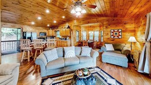 A cabin interior with a wooden ceiling, a dining area, a living room with a sofa and ottoman, and a kitchen with wooden cabinets.