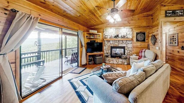 A wooden cabin interior with a stone fireplace, a television, a sofa, and a balcony with a view.