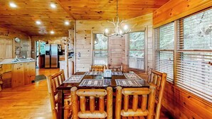 A wooden cabin interior with a dining table, chairs, a chandelier, and a kitchen area.