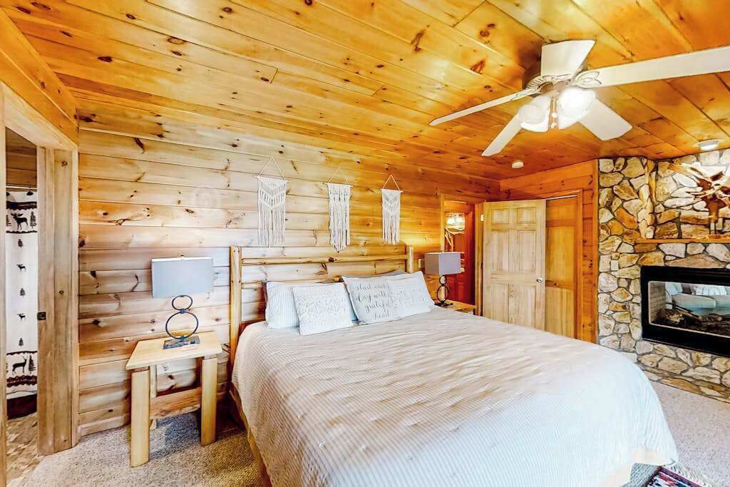 Une chambre à coucher dans une cabane en bois, avec un lit, des tables de chevet, un ventilateur de plafond, un mur en pierre et une cheminée.