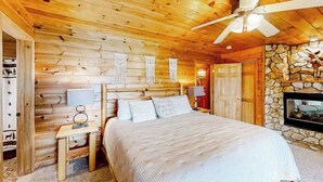 A wooden cabin bedroom with a bed, nightstands, a ceiling fan, a stone wall, and a fireplace.