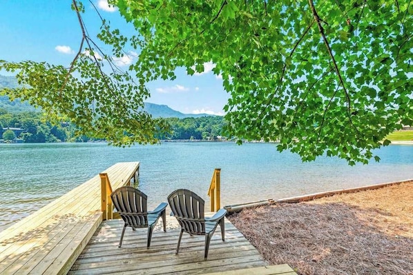 Eine Holzstege mit zwei Stühlen, der auf einen stillen See mit umliegenden Bäumen blickt.
