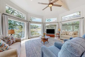 Ein geräumiges Wohnzimmer mit Deckenventilator, großen Fenstern, Fernseher und einer Couch.