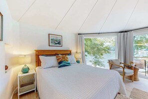 Ein Schlafzimmer mit einem großen Bett, einem hölzernen Kopfteil, einem gerahmten Bild, einem Fenster mit Blick auf Grünflächen und einem Stuhl am Fenster.