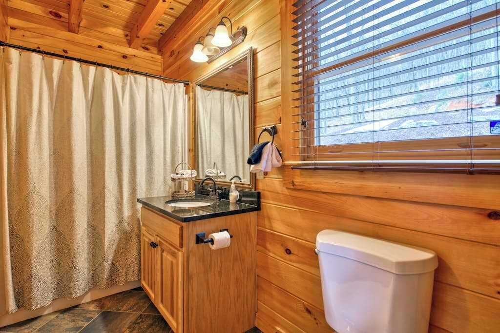 Baño de cabaña de madera con lavamanos, espejo, inodoro y ventana con persianas.