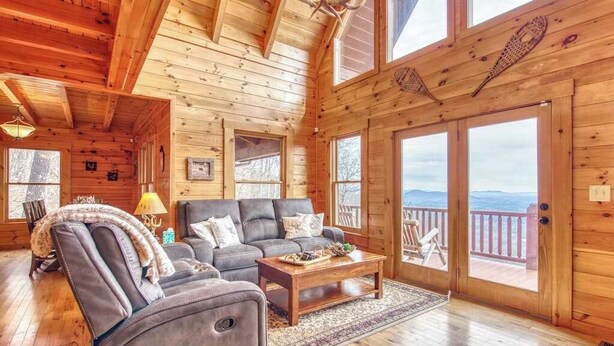 A log cabin interior with a living room, sofa, coffee table, and a view through the door. - Rustic mountain-view home with deck, fire pit, hot tub, fireplace, & game room (Lake Lure)