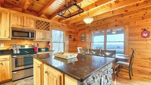 A wooden cabin interior with a kitchen, stone island, and dining area. - Rustic mountain-view home with deck, fire pit, hot tub, fireplace, & game room (Lake Lure)