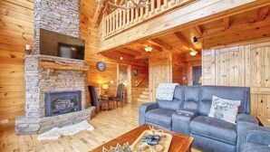 A log cabin interior with a stone fireplace, a leather sofa, and a wooden dining area. - Rustic mountain-view home with deck, fire pit, hot tub, fireplace, & game room (Lake Lure)
