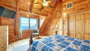 A wooden cabin bedroom with a bed, a chair, a ceiling fan, and a large window. - Rustic mountain-view home with deck, fire pit, hot tub, fireplace, & game room (Lake Lure)