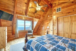 Ein Schlafzimmer in einer Holzhütte mit einem Bett, einem Sessel, einem Deckenventilator und einem großen Fenster.