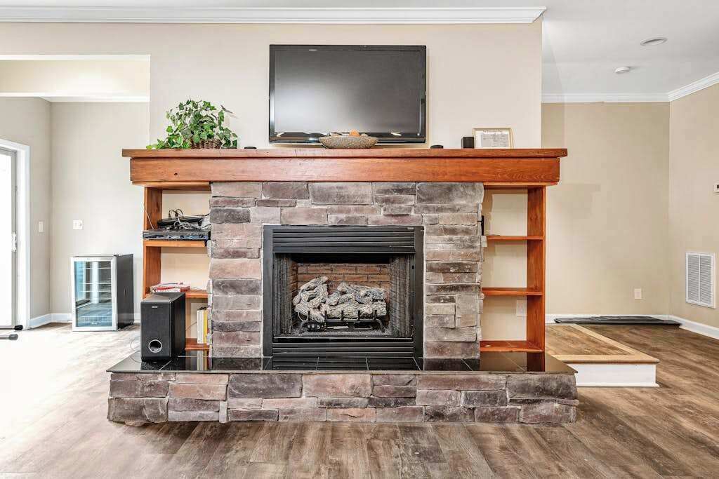 A living room with a stone fireplace, a flat-screen TV above it, and a bookshelf to the right.