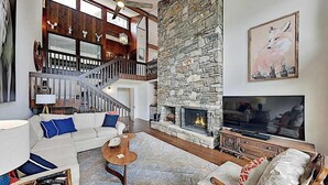 A spacious living room with a stone fireplace, a staircase, and a large TV. - Mountain gem w/pool,golf, hot tub, tennis, beach, wellness center - dog-friendly (Lake Lure)