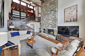 A spacious living room with a stone fireplace, a staircase, and a large TV. - Mountain gem w/pool,golf, hot tub, tennis, beach, wellness center - dog-friendly (Lake Lure)