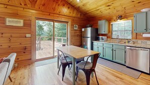 A wooden cabin interior with a dining table, chairs, a kitchen area, and a sliding glass door leading to an outdoor deck. - Happy Top Hideaway (Bryson City)