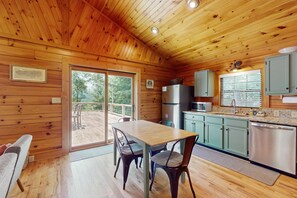 A wooden cabin interior with a dining table, chairs, a kitchen area, and a sliding glass door leading to an outdoor deck. - Happy Top Hideaway (Bryson City)