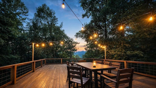 A wooden deck with a table and chairs, surrounded by trees and string lights. - Happy Top Hideaway (Bryson City)