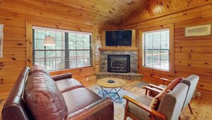 A log cabin interior with a fireplace, leather sofa, wooden chairs, and large windows. - Happy Top Hideaway (Bryson City)