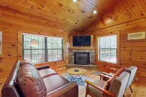 A log cabin interior with a fireplace, leather sofa, wooden chairs, and large windows. - Happy Top Hideaway (Bryson City)