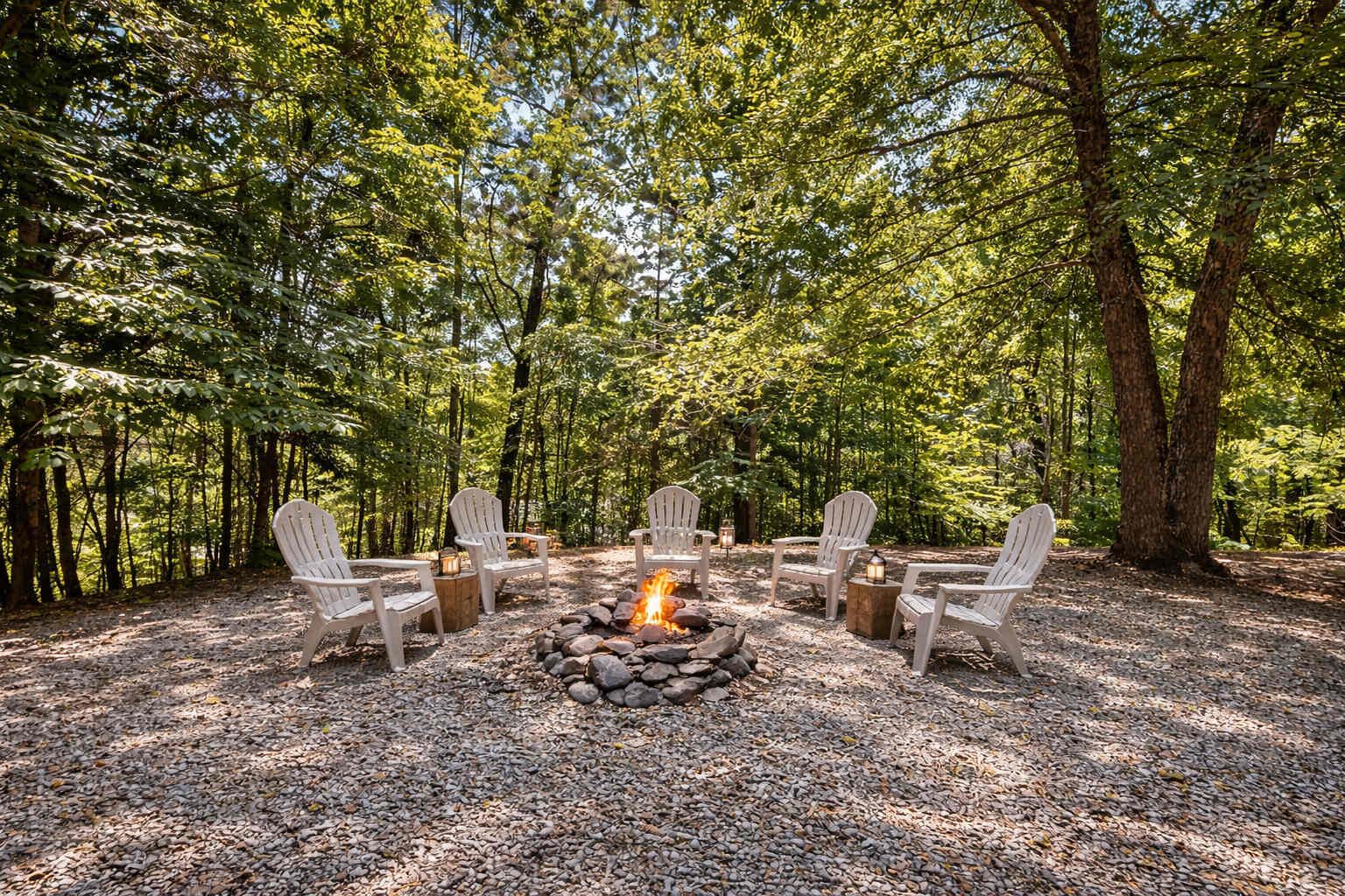 A forested area with a fire pit and white Adirondack chairs arranged around it.