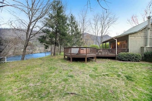 A house with a deck overlooking a body of water, surrounded by trees and a hilly landscape.