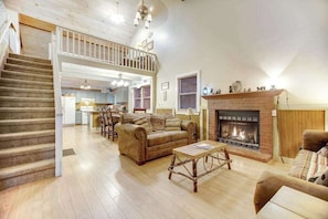 A spacious living room with a fireplace, a staircase, and a kitchen in the background.