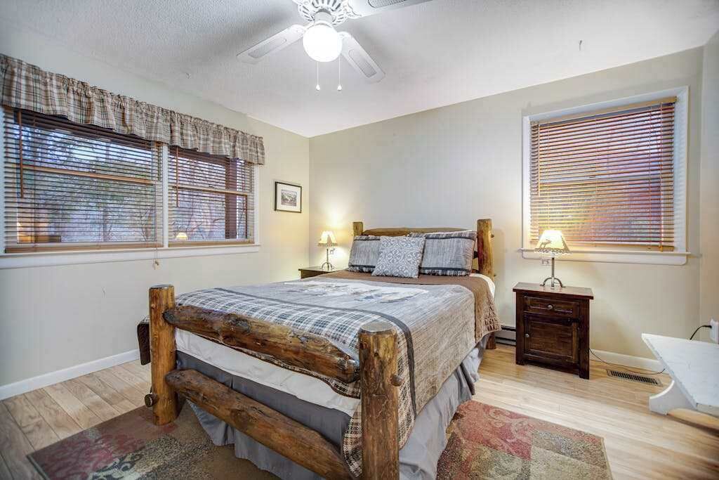 Quarto com cama de madeira, duas mesas de cabeceira, ventilador de teto e janela com persianas.