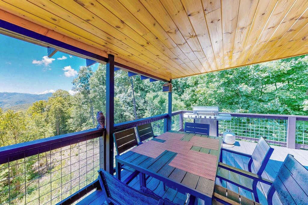 A covered outdoor seating area with a table and chairs, overlooking a forested landscape.