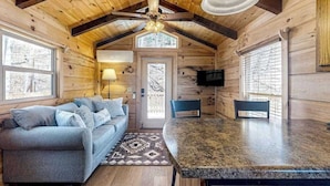 A log cabin interior with a sofa, dining table, chairs, a ceiling fan, and a TV mounted on the wall.