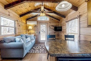 Ein Holzblockhaus-Interieur mit einer Couch, einem Esstisch, Stühlen, einem Deckenventilator und einem an der Wand befestigten Fernseher.