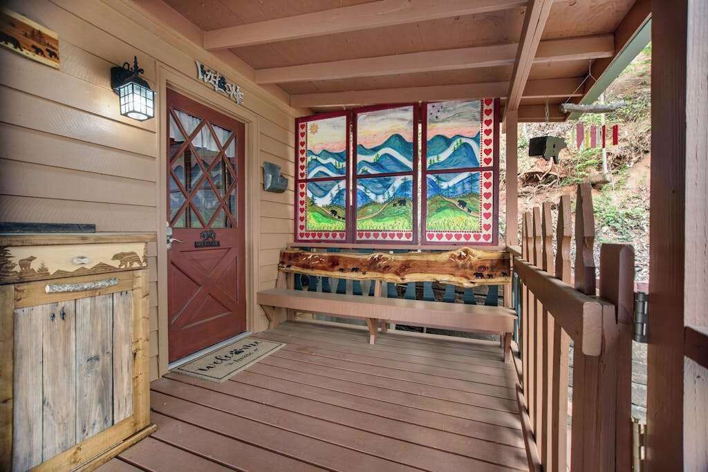 A wooden porch with a bench, a red door, and a window with a landscape painting.