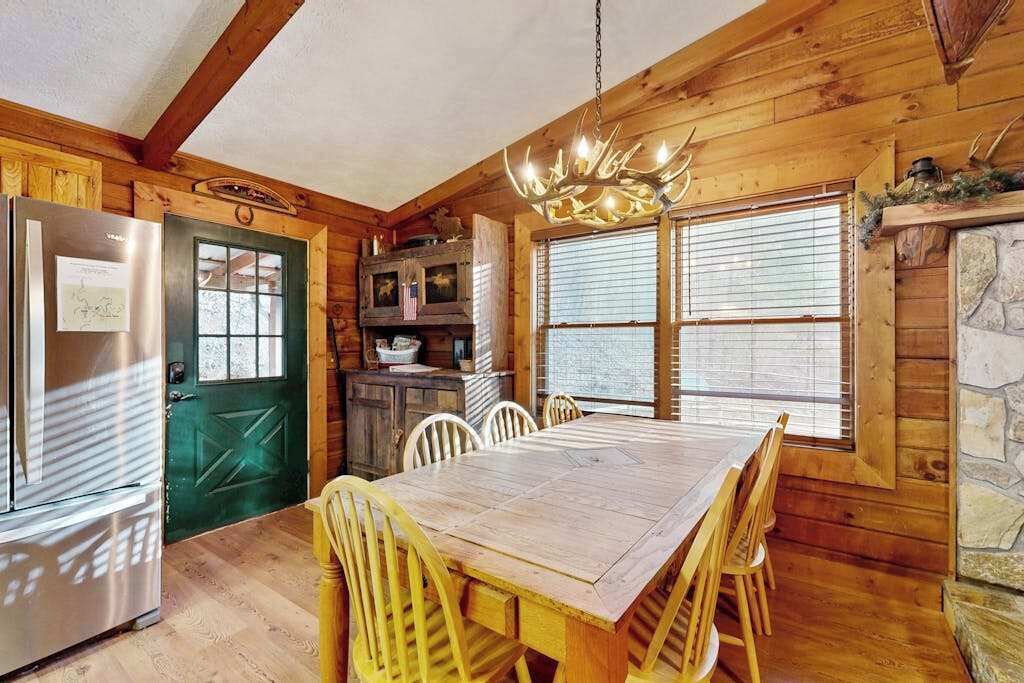 Interior de una cabaña de madera con una mesa de comedor, sillas, un refrigerador, una puerta verde y una chimenea de piedra.