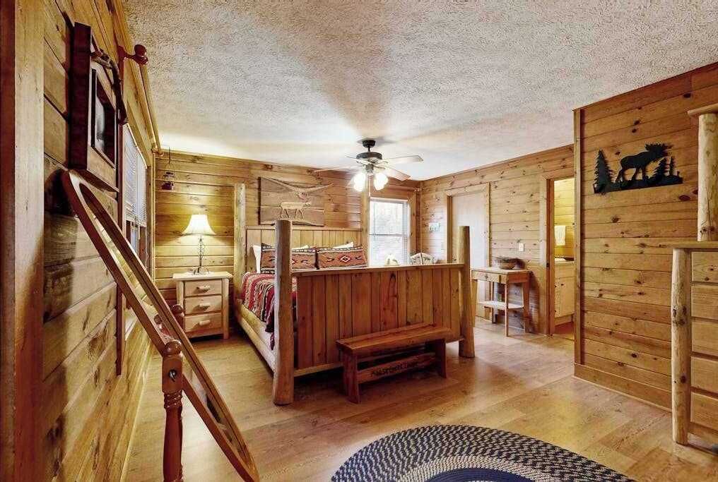 A log cabin interior with a staircase, a bed, a desk, and a ceiling fan.
