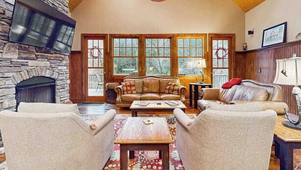 A spacious living room with a stone fireplace, wooden ceiling, and large windows. - Tuckaway Retreat (Sylva)