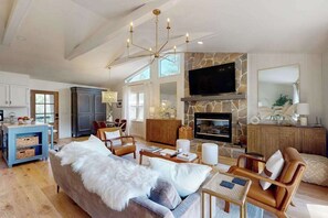 A spacious living room with a stone fireplace, a television mounted above it, and a large window providing natural light. - Cashmere Cottage (Cashiers)