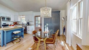Ett modernt kök med en central köksö, en matplats med ett glasbord och ett stort fönster som släpper in naturligt ljus.