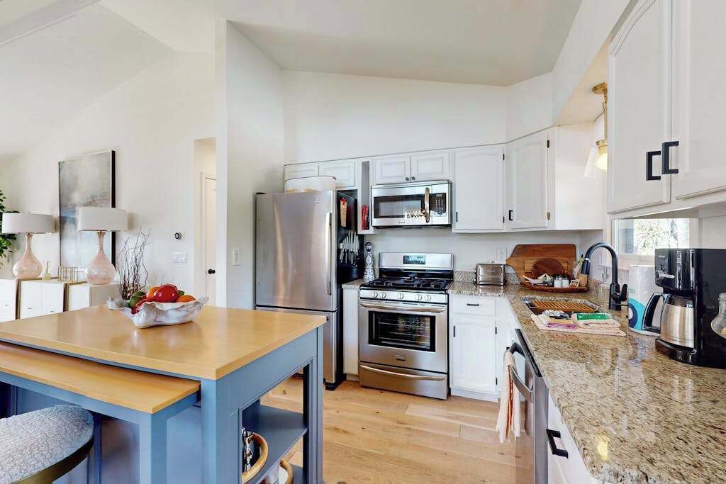 A modern kitchen with white cabinets, stainless steel appliances, a granite countertop, and a wooden dining table.
