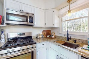 A kitchen with white cabinets, a stainless steel stove, a microwave, a sink, and a window with a view of trees. - Cashmere Cottage (Cashiers)