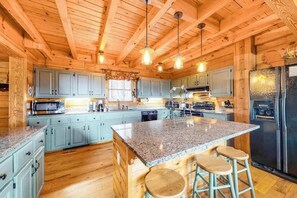A wooden cabin interior with a kitchen featuring granite countertops, bar stools, and a refrigerator. - Secluded cabin with mountain views, fire pit & billiards (Sylva)